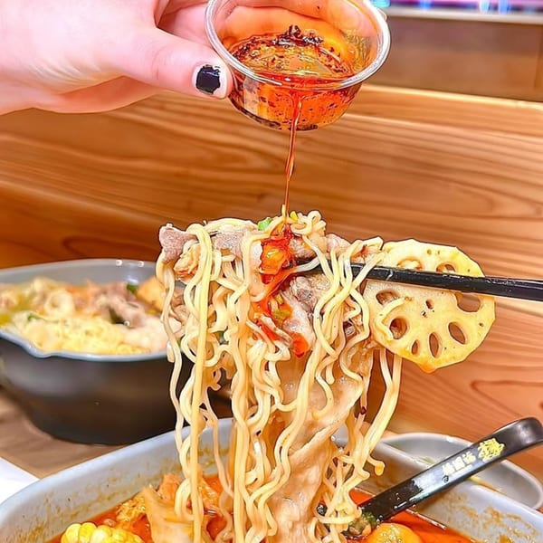 A hand pours chili oil over a spoonful of YGF Malatang noodles, meat, and lotus root.