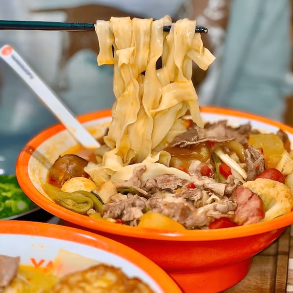 Chopsticks lifting wide noodles from a bowl of YGF Malatang with meat and vegetables.