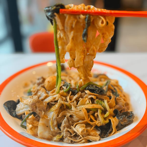 Chopsticks lifting saucy YGF Malatang noodles with meat, vegetables, and mushrooms from a bowl.