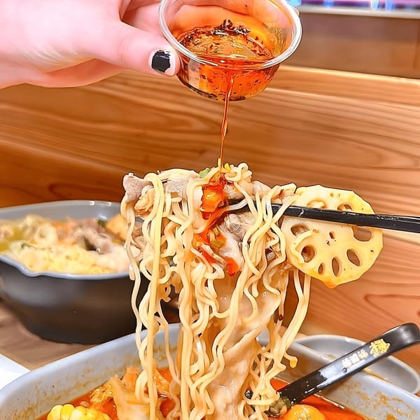 A hand pours chili oil over YGF Malatang noodles, meat, and lotus root held by chopsticks.