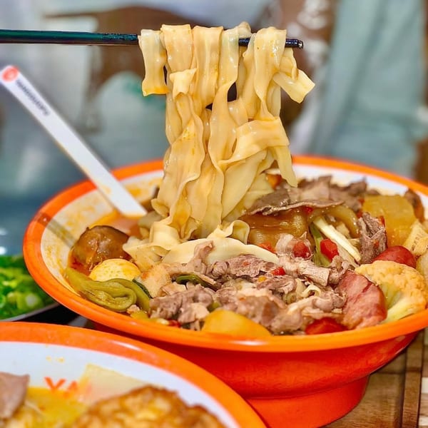 Chopsticks lift wide noodles from a bowl of YGF Malatang with meat, vegetables, and broth.